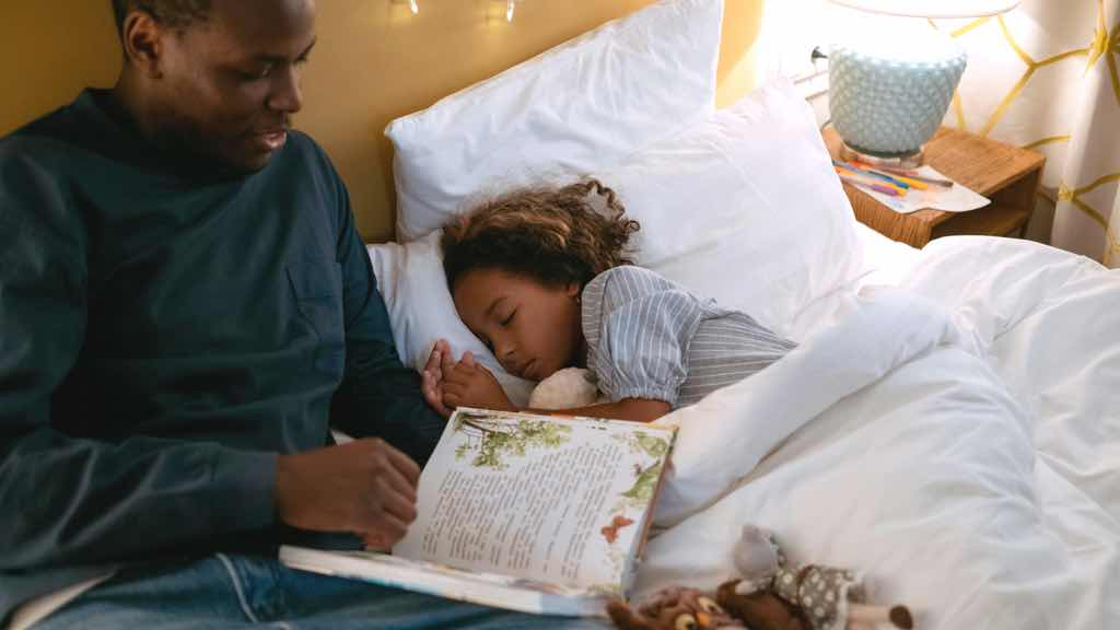Father reading bedtime story for his daughter while lying down on the bed; image by cottonbro studio.